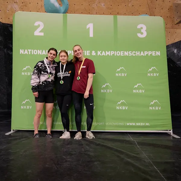 Drie jonge vrouwen met medailles staan op een podium voor een groene achtergrond waarop ‘NKBV’ en ‘NATIONALE KLIMKAMPIOENSCHAPPEN’ staat geschreven, terwijl ze hun overwinning vieren tijdens een klimkampioenschap.